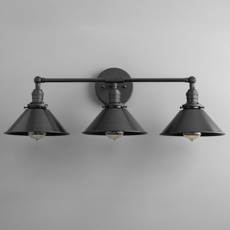 May include: Black three-light vanity light fixture with black metal shades. The fixture has a simple, industrial design.