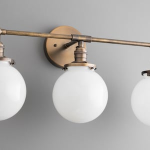 May include: A three-light vanity fixture with a brass finish and white glass globes. The fixture has a long, straight arm with adjustable arms for each light.