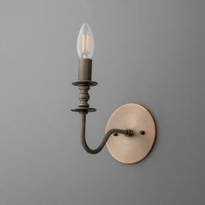 May include: A vintage-style wall sconce with a brass finish. The sconce features a curved arm, a round backplate, and a decorative candle-style holder with a clear, energy-efficient LED bulb. The fixture is designed for indoor use.