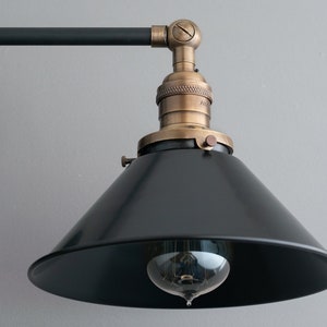 May include: Black metal wall sconce with two adjustable arms and black cone shades. Each shade has a brass socket and a vintage-style light bulb.
