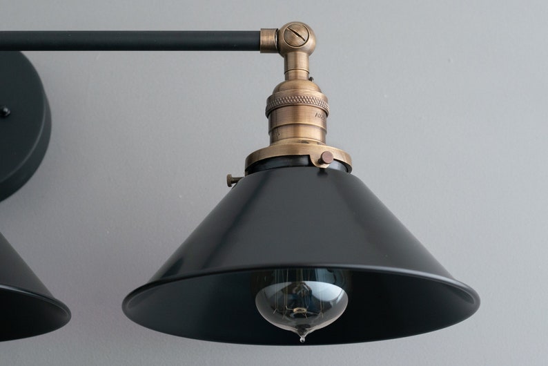 May include: Black metal wall sconce with two adjustable arms and black cone shades. Each shade has a brass socket and a vintage-style light bulb.