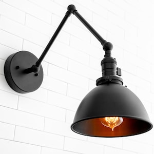 May include: Black industrial wall sconce with a round, black metal shade and an adjustable arm. The sconce is mounted on a white brick wall and has a warm-toned light bulb.