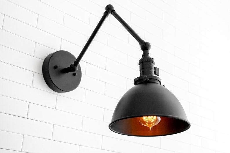 May include: Black industrial wall sconce with a round, black metal shade and an adjustable arm. The sconce is mounted on a white brick wall and has a warm-toned light bulb.