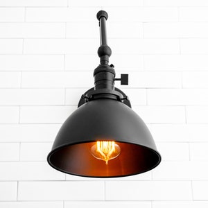 May include: Black metal wall sconce with a dome-shaped shade and an exposed bulb. The sconce is mounted on a white brick wall.