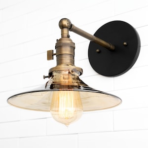 May include: A brass wall sconce with a smoked glass shade and a black circular backplate. The sconce has a single bulb and is mounted on a white brick wall.