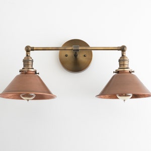 May include: A vintage-style wall sconce with two lights. The fixture features a round brass backplate, a horizontal brass bar, and two copper-colored cone-shaped shades. Each shade houses a visible light bulb, creating a warm, inviting glow. The fixture is mounted on a white wall.
