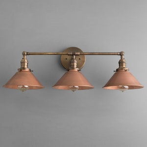 May include: A three-light vanity fixture with a vintage industrial design. It features three copper-colored cone-shaped shades, each housing a lightbulb. The fixture is mounted on a brass-toned metal frame and backplate, creating a warm, inviting aesthetic.