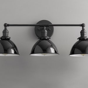 Puede incluir: Aplique de ba&ntilde;o negro de tres luces con pantallas de metal negro. La luminaria tiene un dise&ntilde;o de barra recta con tres luces espaciadas uniformemente.