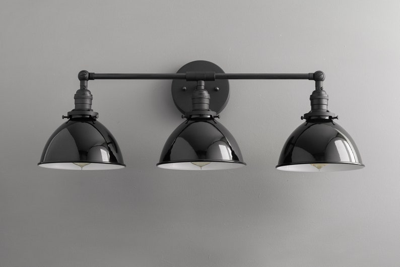 Puede incluir: Aplique de ba&ntilde;o negro de tres luces con pantallas de metal negro. La luminaria tiene un dise&ntilde;o de barra recta con tres luces espaciadas uniformemente.