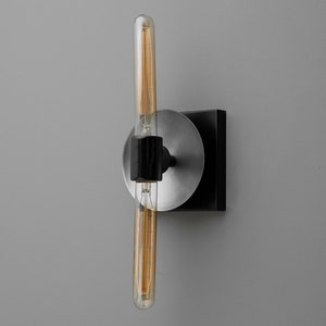 May include: A wall-mounted light fixture featuring a long, cylindrical Edison-style light bulb with visible filaments. The bulb is held by a black central bracket attached to a circular brushed silver plate and a square black base mount.
