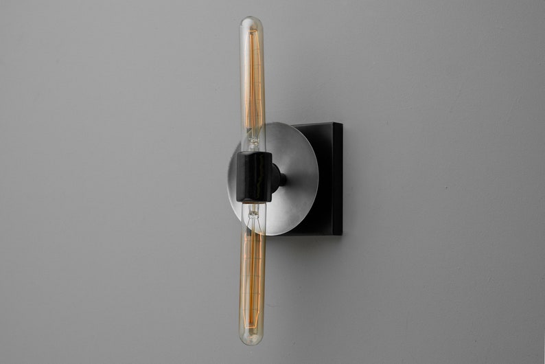 May include: A wall-mounted light fixture featuring a long, cylindrical Edison-style light bulb with visible filaments. The bulb is held by a black central bracket attached to a circular brushed silver plate and a square black base mount.