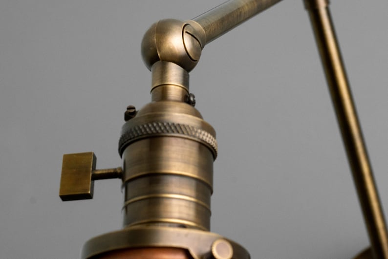 K&ouml;nnte beinhalten: Nahaufnahme einer antiken Schreibtischlampe aus Messing mit verstellbarem Arm und Schirm. Die Lampe hat ein Vintage-Design mit einem quadratischen Schalter und einer detaillierten Lampenfassung. Die Oberfl&auml;che ist in einem warmen, gealterten Messington gehalten.