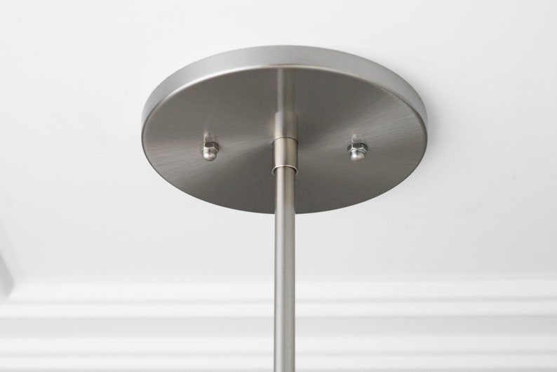 May include: Close-up of a brushed nickel ceiling mount for a pendant light fixture. The mount has two screws and a center hole for the hanging rod.
