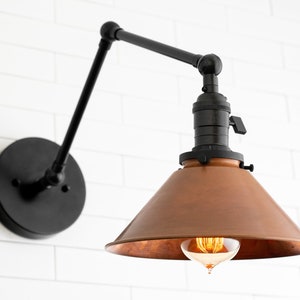 K&ouml;nnte beinhalten: Eine schwarze Wandleuchte mit kupferfarbenem Schirm. Die Lampe hat einen verstellbaren Arm und einen runden Sockel. Die Gl&uuml;hbirne ist sichtbar. Die Lampe ist an einer wei&szlig;en Ziegelmauer befestigt.