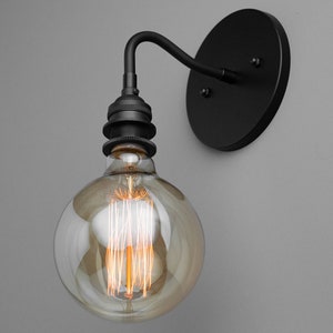 K&ouml;nnte beinhalten: Eine schwarze Wandleuchte mit einer runden R&uuml;ckplatte und einem gebogenen Arm. Eine klare Glaskugel-Gl&uuml;hbirne mit einem Filament ist am Arm befestigt.