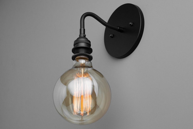 K&ouml;nnte beinhalten: Eine schwarze Wandleuchte mit einer runden R&uuml;ckplatte und einem gebogenen Arm. Eine klare Glaskugel-Gl&uuml;hbirne mit einem Filament ist am Arm befestigt.