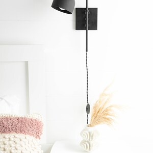 Op de afbeelding: Zwarte wandlamp met een zwart snoer en een zwarte kap. De lamp is gemonteerd aan een witte muur naast een wit nachtkastje met een witte vaas en gedroogd pampasgras.