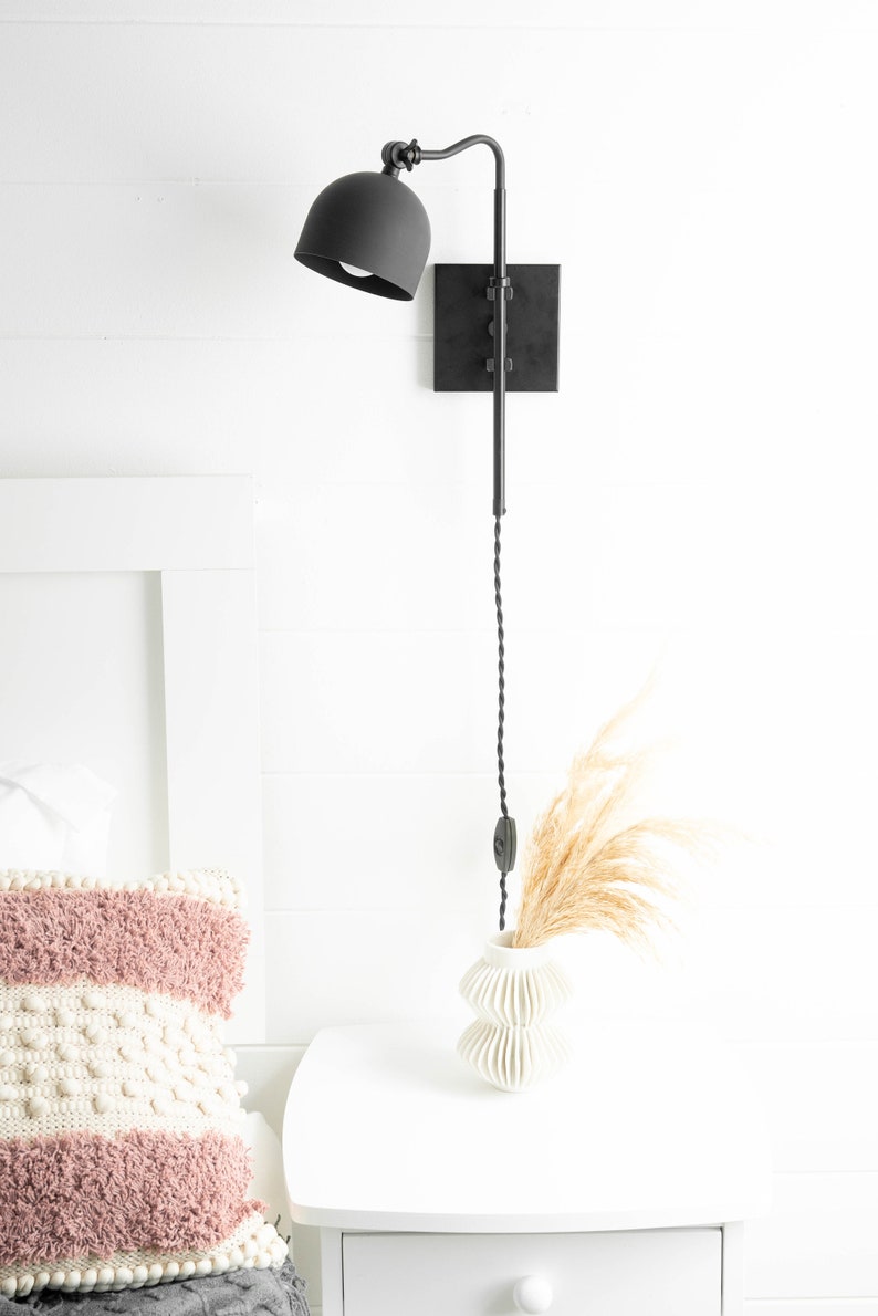 Op de afbeelding: Zwarte wandlamp met een zwart snoer en een zwarte kap. De lamp is gemonteerd aan een witte muur naast een wit nachtkastje met een witte vaas en gedroogd pampasgras.