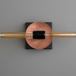 May include: A wall-mounted light fixture featuring a long, horizontal tubular filament bulb centered on a black cylindrical socket. The socket is mounted onto a circular copper-toned plate, which is attached to a square black base plate.