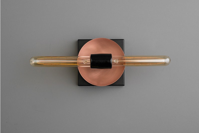 May include: A wall-mounted light fixture featuring a long, horizontal tubular filament bulb centered on a black cylindrical socket. The socket is mounted onto a circular copper-toned plate, which is attached to a square black base plate.