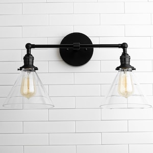 May include: Black, two-light vanity fixture with clear glass shades. The fixture has a round backplate and a horizontal bar with two downward-facing lights. The bulbs are visible inside the glass shades. The fixture is mounted on a white, tiled wall.