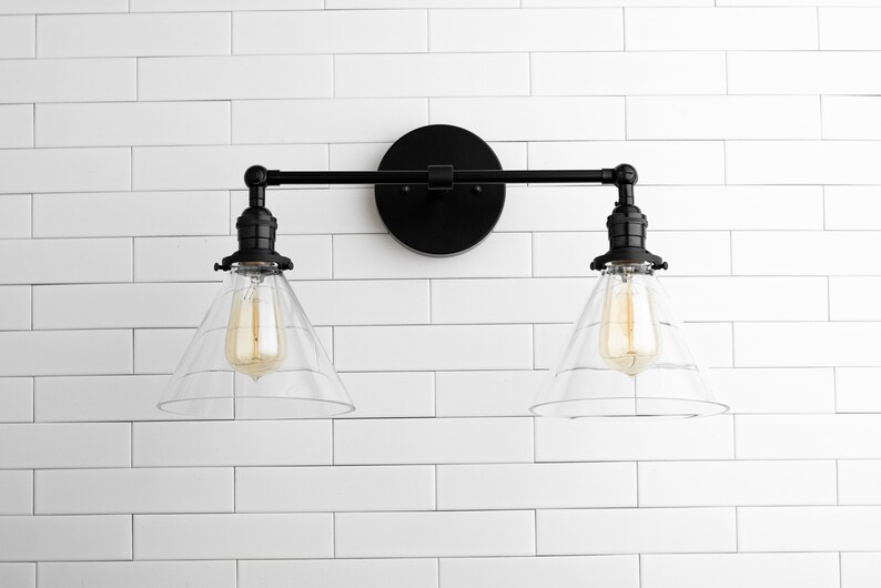 May include: Black, two-light vanity fixture with clear glass shades. The fixture has a round backplate and a horizontal bar with two downward-facing lights. The bulbs are visible inside the glass shades. The fixture is mounted on a white, tiled wall.