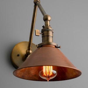 K&ouml;nnte beinhalten: Vintage Wandleuchte mit kupferfarbenem, konischem Schirm und freiliegender Gl&uuml;hbirne. Die Leuchte verf&uuml;gt &uuml;ber messingfarbene, verstellbare Arme und eine runde Wandplatte. Die Leuchte ist f&uuml;r die Wandmontage konzipiert.