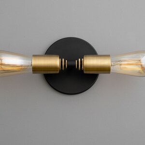 Op de afbeelding: Een zwarte en gouden wandlamp met twee gloeilampen. De lamp heeft een ronde zwarte basis en twee gouden armen die naar de zijkanten uitsteken. De gloeilampen zijn van helder glas en hebben een warmwitte gloed.