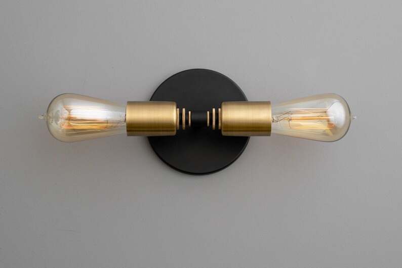 Op de afbeelding: Een zwarte en gouden wandlamp met twee gloeilampen. De lamp heeft een ronde zwarte basis en twee gouden armen die naar de zijkanten uitsteken. De gloeilampen zijn van helder glas en hebben een warmwitte gloed.