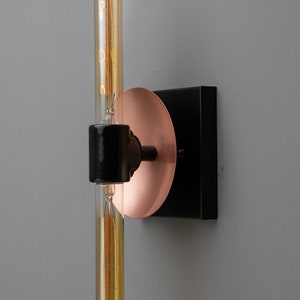 May include: A wall-mounted light fixture featuring a black square base, a circular copper-colored plate, and a central black cylindrical socket holding a long, vertical, tubular Edison-style light bulb with visible internal glowing filaments.