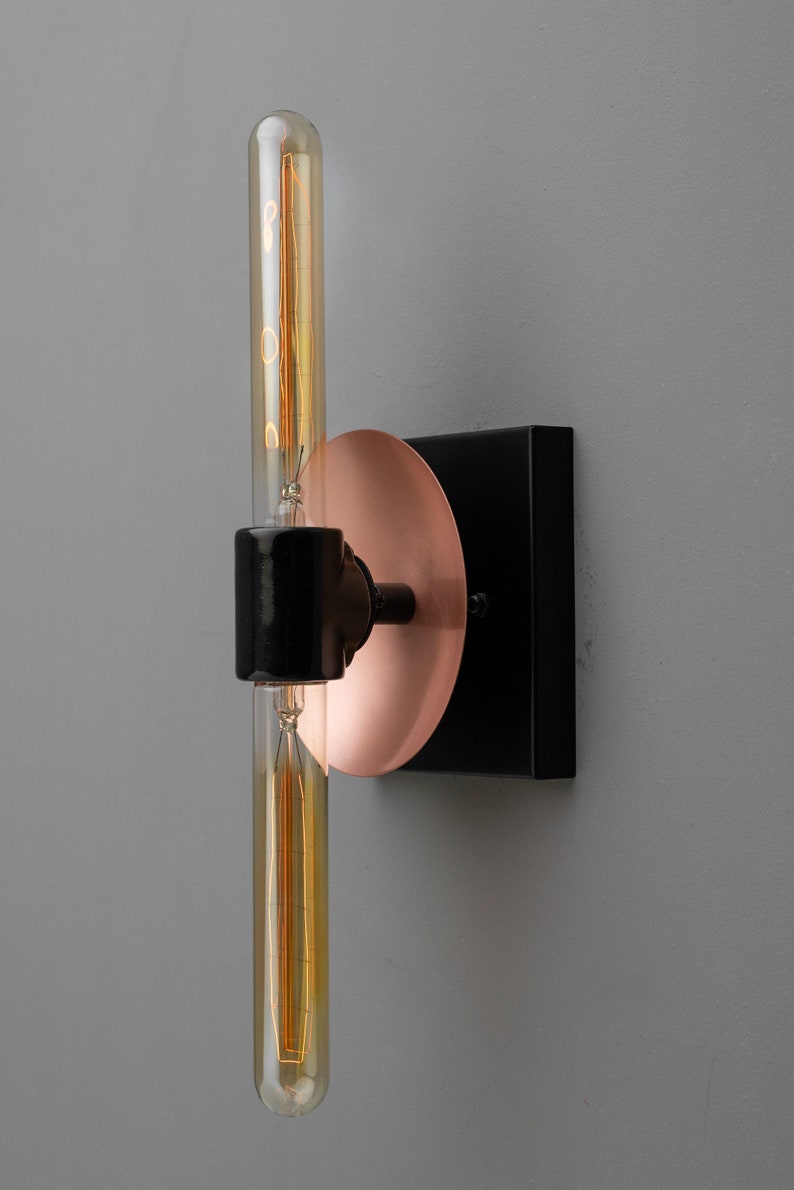May include: A wall-mounted light fixture featuring a black square base, a circular copper-colored plate, and a central black cylindrical socket holding a long, vertical, tubular Edison-style light bulb with visible internal glowing filaments.