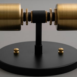 Op de afbeelding: Een zwarte metalen wandlamp met twee gouden cilindrische lampenkappen. De lamp heeft een ronde basis met twee gouden schroeven.