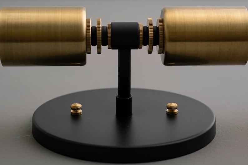 Op de afbeelding: Een zwarte metalen wandlamp met twee gouden cilindrische lampenkappen. De lamp heeft een ronde basis met twee gouden schroeven.