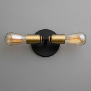 Op de afbeelding: Een zwarte en gouden wandlamp met twee lampen. De lamp heeft een ronde zwarte basis en twee gouden armen die naar de zijkanten uitsteken. De lampen zijn van helder glas met een zichtbaar filament.