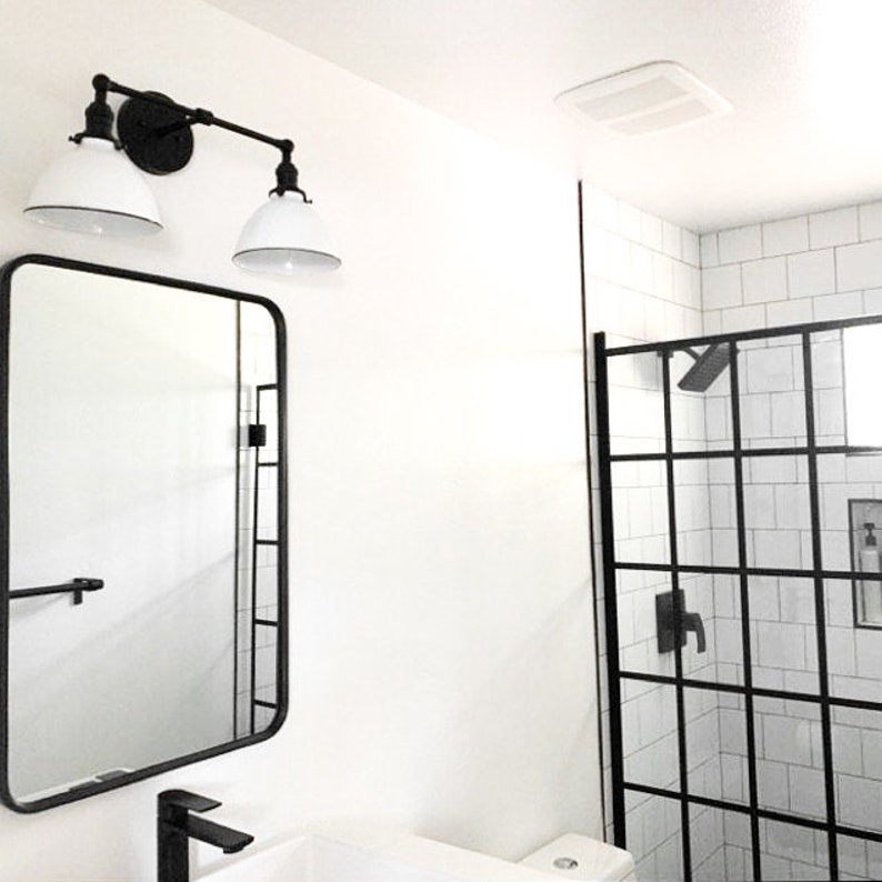 May include: A modern bathroom scene featuring a black-framed mirror, a black faucet, and a two-light fixture with white shades. The shower has a black grid frame. The walls are white, creating a clean, minimalist aesthetic.