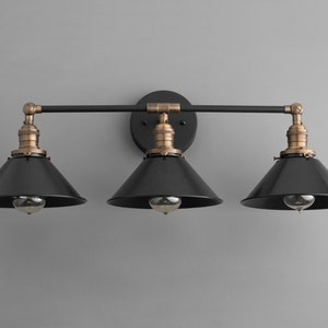 May include: A black three-light vanity light fixture with brass accents and black metal shades. The fixture has a straight bar design with three lights spaced evenly apart.