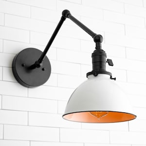 May include: A white and black wall-mounted light fixture with a white shade and a black metal arm. The shade has a gold interior and the light is on.