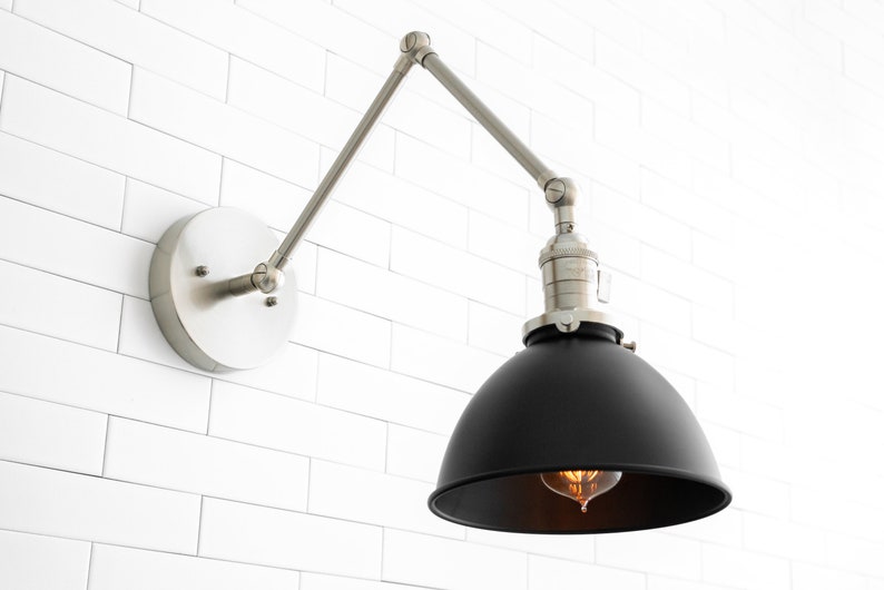 May include: A black metal wall sconce with a silver adjustable arm. The sconce has a black metal shade and a clear light bulb.
