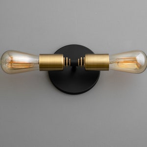 Op de afbeelding: Een zwarte wandlamp met twee gouden armen en heldere glazen bollen. De lamp heeft een ronde zwarte basis.