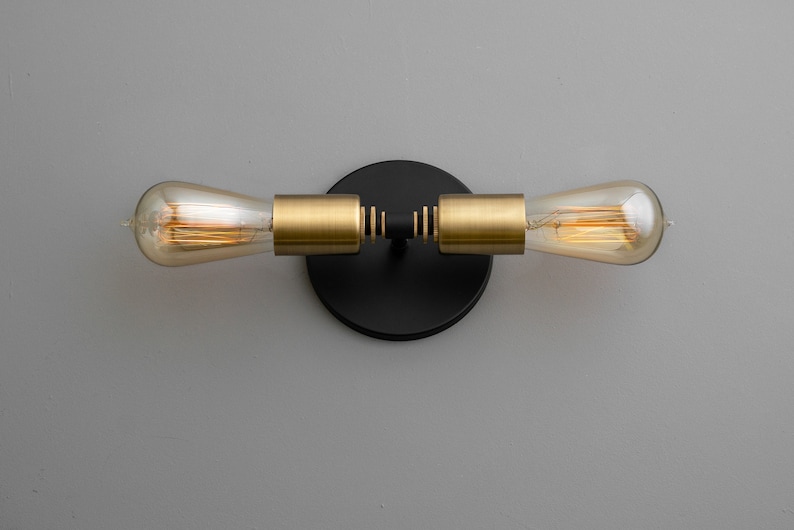 Op de afbeelding: Een zwarte wandlamp met twee gouden armen en heldere glazen bollen. De lamp heeft een ronde zwarte basis.