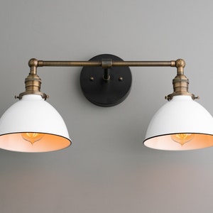 May include: A two-light vanity fixture with white, dome-shaped shades and a black backplate. The fixture has a bronze-colored arm and accents. The interior of the shades is a warm, light color. The fixture is mounted on a gray wall.