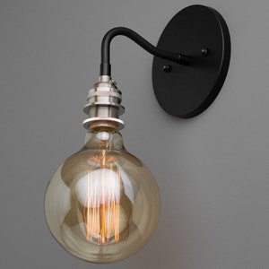 K&ouml;nnte beinhalten: Eine schwarze Wandleuchte mit einer runden schwarzen R&uuml;ckplatte und einem gebogenen Arm. Eine klare Gl&uuml;hbirne aus Glas mit einem Filament ist am Arm befestigt.