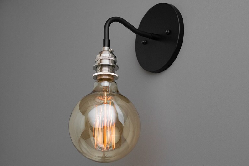 K&ouml;nnte beinhalten: Eine schwarze Wandleuchte mit einer runden schwarzen R&uuml;ckplatte und einem gebogenen Arm. Eine klare Gl&uuml;hbirne aus Glas mit einem Filament ist am Arm befestigt.