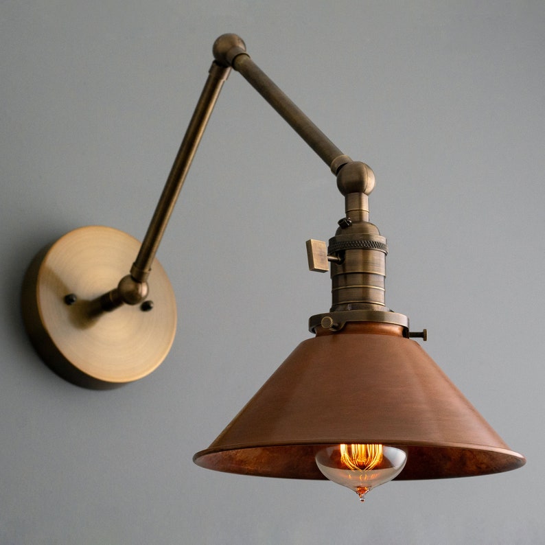 K&ouml;nnte beinhalten: Vintage Wandleuchte mit Bronze-Finish. Die Leuchte verf&uuml;gt &uuml;ber einen verstellbaren Arm und einen konischen Kupferschirm. Die Gl&uuml;hbirne ist sichtbar und strahlt ein warmes Licht aus. Die Leuchte ist auf einer runden Bronze-Basis montiert.