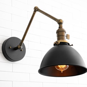 May include: A black and brass wall-mounted light fixture with a black metal shade and a brass arm. The light is on and the bulb is visible.