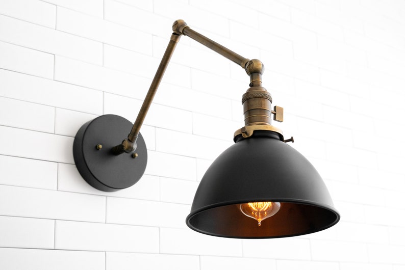 May include: A black and brass wall-mounted light fixture with a black metal shade and a brass arm. The light is on and the bulb is visible.