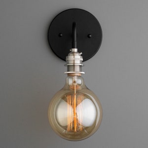 K&ouml;nnte beinhalten: Eine schwarze Wandleuchte aus Metall mit einer runden R&uuml;ckplatte und einer einzelnen Lampenfassung. Die Leuchte ist an einer grauen Wand befestigt und verf&uuml;gt &uuml;ber eine klare Glaskugellampe mit einem Filament.
