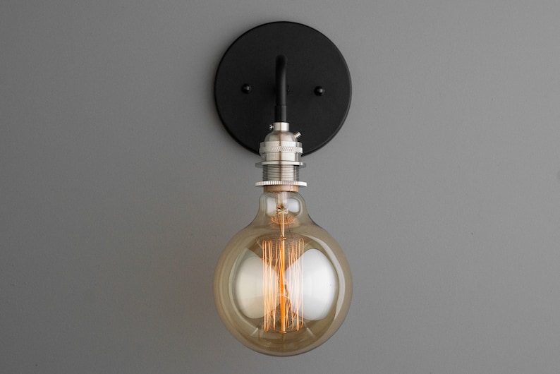 K&ouml;nnte beinhalten: Eine schwarze Wandleuchte aus Metall mit einer runden R&uuml;ckplatte und einer einzelnen Lampenfassung. Die Leuchte ist an einer grauen Wand befestigt und verf&uuml;gt &uuml;ber eine klare Glaskugellampe mit einem Filament.