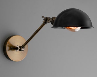 May include: A wall-mounted lamp featuring a matte black dome-shaped shade and an adjustable arm with a brass finish. The fixture includes a circular brass wall plate and a single exposed glowing light bulb visible beneath the shade.