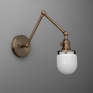 May include: A wall-mounted, adjustable brass-toned lamp with a white, teardrop-shaped glass shade. The lamp features a round base and an articulated arm for directional lighting. The design is reminiscent of vintage industrial lighting.